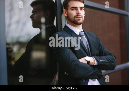 Portrait of confident businessman standing with his arms crossed. Man in formal wear leaning to glass outdoors and looking at camera. Stock Photo