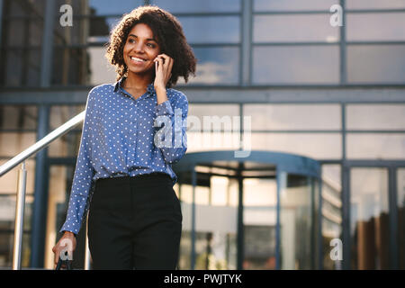 Smiling businesswoman talking over mobile phone while walking outside office building. Woman in formalwear walking outdoors and talking on phone. Stock Photo