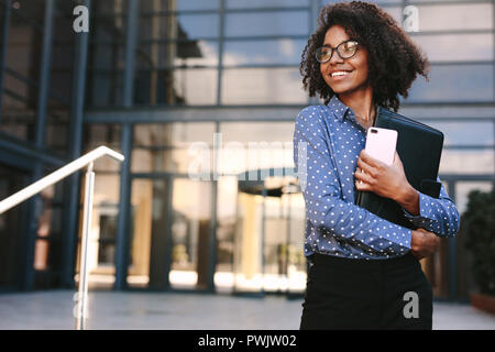 Woman with mobile phone and file standing outdoors and looking away. Business woman standing outside a office building. Stock Photo