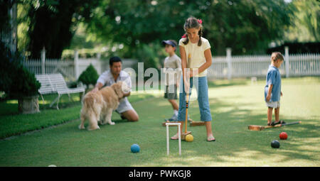 Girl playing croquet on a grassy lawn with her father and two younger brothers. Stock Photo