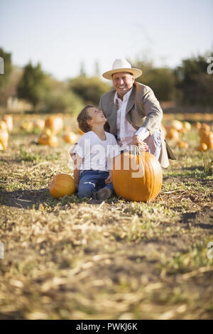 Portrait of a smiling mid-adult man holding a pumpkin while sitting with his son in a pumpkin patch, Stock Photo