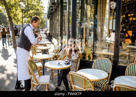 PARIS, FRANCE - August 31, 2018: Waiter serving coffee to a young woman client sitting at the traditional french cafe outdoors in Paris Stock Photo