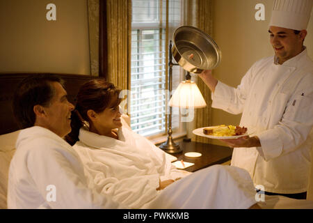 Young adult male chef presenting room service to a mid-adult couple in their hotel room. Stock Photo