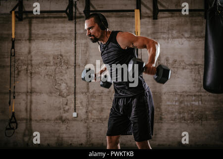 Young muscular man doing hard exercise with dumbbells for shoulders on cross fit training at the gym. Stock Photo