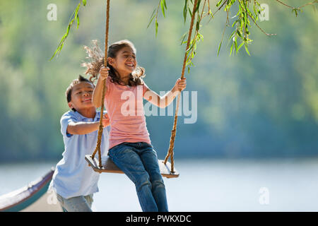 Happy young boy pushing a young girl on a tree swing. Stock Photo