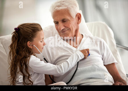 Elderly man receiving a hospital visit from his granddaughter,  who is playing with a stethoscope. Stock Photo