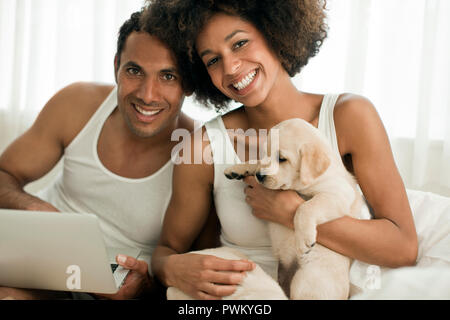 Happy couple holding laptop and Labrador puppy in bed together. Stock Photo