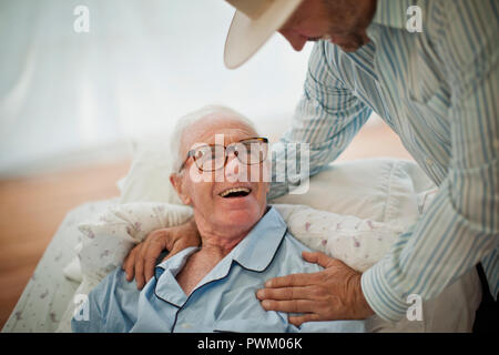 Smiling senior man being comforted by his adult son while lying in bed in his pajamas. Stock Photo