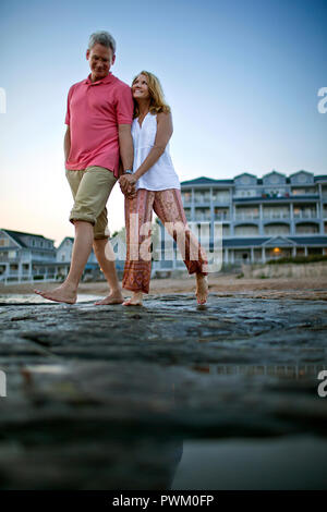 Mature couple holding hands and smiling as they walk barefoot on the beach at sunset. Stock Photo