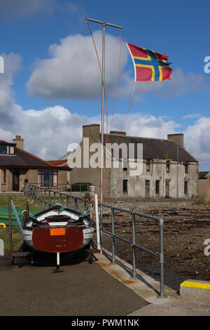 A very tattered flag of Orkney, Scotland, flies in a strong wind in from the pier in St. Mary's. Bright blue sky, white clouds, boat, building, shore. Stock Photo