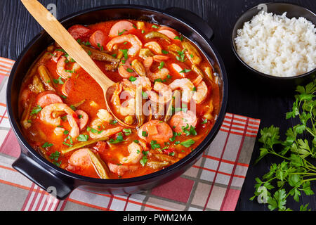 Delicious gumbo with prawns, sausage in a dutch oven on a black table with bowl of white rice, horizontal view from above, close-up Stock Photo