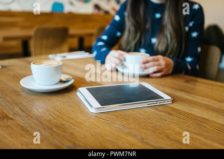 Cappuccino coffee in a mug on the table next to the tablet. The girl is drinking coffee in the background. Relaxing or meeting in a cafe. The girl is waiting for someone else. Stock Photo
