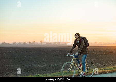 Female commuter riding a bike out of town in rural area. Young woman riding bike at sunset Stock Photo