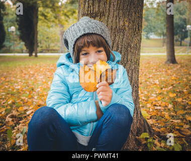 Portrait of six years old caucasian girl in autumn. She's wearing a light blue jacket and a grey wool cap and holds in her hands many fall leaves Stock Photo