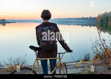 Female cyclist enjoying beautiful blue hour scene by the lake. Woman stands with bike and looks at beautiful lake and sunset Stock Photo