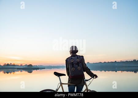 Female cyclist enjoying beautiful blue hour scene by the lake. Woman stands with bike and looks at beautiful lake and sunset Stock Photo