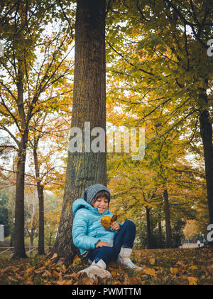 Portrait of six years old caucasian girl in autumn. She's wearing a light blue jacket and a grey wool cap and holds in her hands many fall leaves Stock Photo