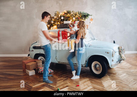 View of lovely daddy giving Christmas present, big red box, for blonde mom and little cute daughter. Family spending time together in home decorated f Stock Photo
