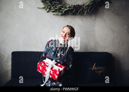 Gorgeous woman holding gift box with red bow. Charming model with blonde hair having perfect make up and red lips. Young woman sitting in living room  Stock Photo