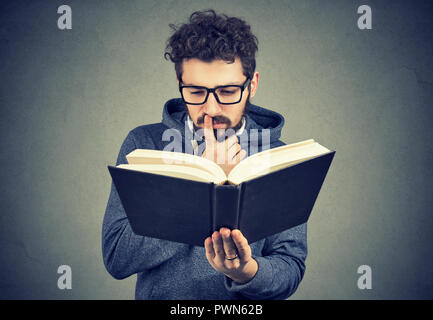 Young man in glasses looking confused while trying to read smart book on gray background