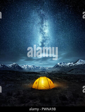 Camping in the wilderness. A pitched tent under the glowing  night sky stars of the milky way with snowy mountains in the background. Nature landscape Stock Photo