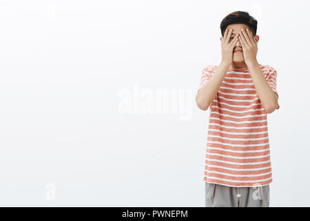 Cute asian young male student eager to see what fate prepared for him, holding hands on face peeking through fingers with one eye anticipating gift or surprise over white background