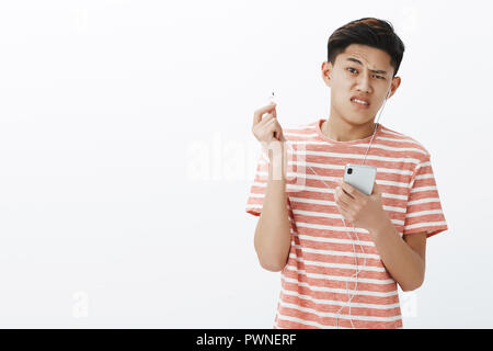 Waist-up shot of displeased cute young asian boy in striped t-shirt taking off broken earbud holding smartphone frowning dissatisfied and bothered with law quality of sound, posing over white wall Stock Photo