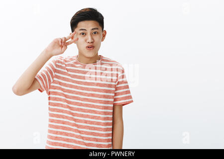 Happy charismatic cute asian guy with short hairstyle smiling folding lips silly making peace sign near face, posing against white background in striped t-shirt, taking photo during family vacation
