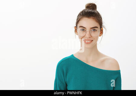 Waist-up shot of smart and creative cute feminine girl in transparent glasses with bun hairstyle, smiling broadly while looking at portrait she draw, standing in gallery over grey wall Stock Photo
