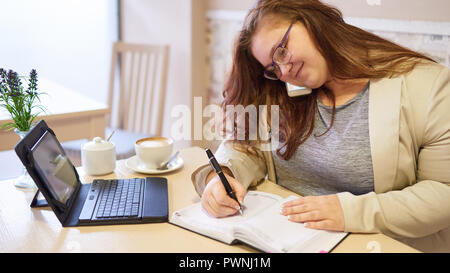 Beautiful plus size business woman with healthy skin and loose brown hair and a subtle smile working in a coffee shop to get ahead before going in to  Stock Photo