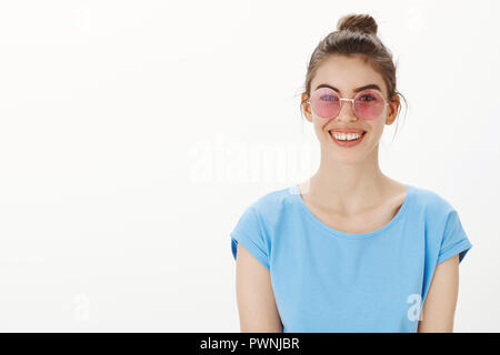 Indoor shot of trendy beautiful woman in blue t-shirt and pink trendy sunglasses, smiling joyfully and carefree while standing over gray background, waiting for friend getting dressed for party Stock Photo