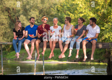 group of young friends enjoying watermelon while sitting on the wooden bridge over the river in beautiful nature Stock Photo