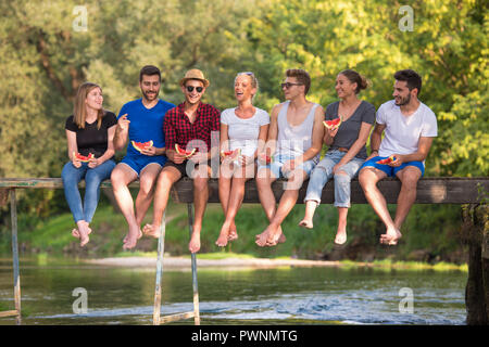 group of young friends enjoying watermelon while sitting on the wooden bridge over the river in beautiful nature Stock Photo