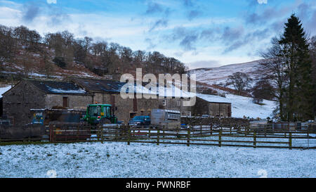 Tractor & trailers by farm buildings & old farmhouse on snowy winter day in scenic, Yorkshire Dales countryside valley - Hubberholme, England, UK. Stock Photo