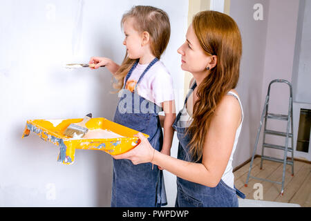 Repair in the apartment. Happy family mother and daughter in aprons paint the wall with white paint. the daughter paints the wall with a brush, and the mother holds the paint cell and observes Stock Photo