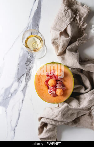 Melon and ham or prosciutto salad served in half of Cantaloupe melon on linen cloth over white marble background with glass of white wine. Flat lay, s Stock Photo