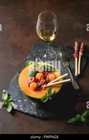 Melon and ham salad served in half of Cantaloupe melon with fresh basil and grissini bread on black slate board over dark brown texture background wit Stock Photo