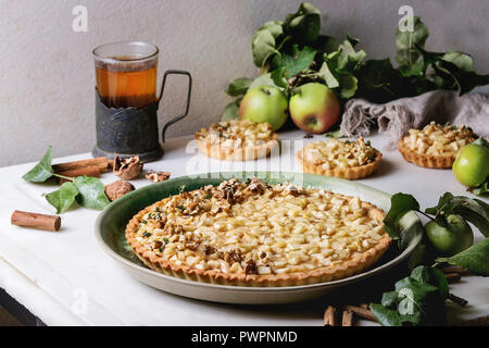 Homemade sweet apple shortbread tart in green ceramic plate, mini tartlets and hot tea in vintage cup holder with cinnamon sticks, walnuts, apples bra Stock Photo