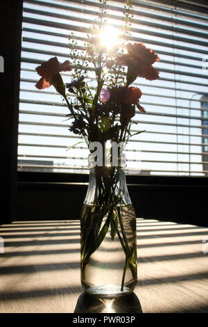 Softly focus of flowers in glass bottle full of water on wooden empty table with horizontal shadows on the table and stunning sunlight coming from a w Stock Photo