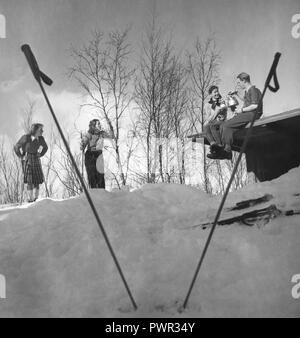 Winter in the 1940s. A young couple is having a cup of coffee while sitting on the roof of their cottage. Their two friends are standing on skis and chatting.  Sweden 1940s. Photo Kristoffersson Ref D115-2 Stock Photo