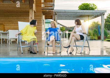 Three mature middle-aged women are having fun and talking, sitting in a lounger by the pool, summer evening. Stock Photo
