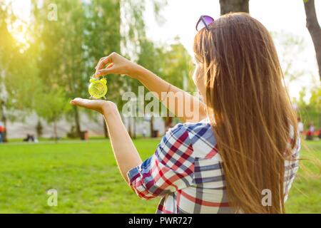 Young woman holding alarm clock in her hand, looking at the time. Background sunny city park, copy space Stock Photo