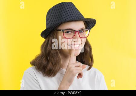 Positive smiling teenage girl 13.14 years old with glasses, a hat shows a sign of silence, holds a finger on her lips, a yellow studio background. Peo Stock Photo
