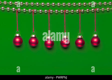 Six small matte red-coloured Christmas ornaments hanging on a row decorated by shiny golden-coloured Christmas beads wreath on green background in stu Stock Photo