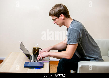 Concentrated young man with glasses working on a laptop in a home office. Type on a keyboard and scrolls text on the display. side view Stock Photo