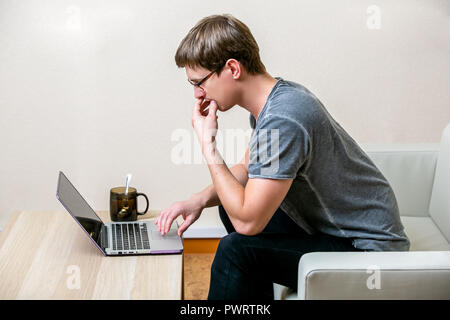 Concentrated young man with glasses working on a laptop in a home office. Pensively scans the text on the display. side view Stock Photo