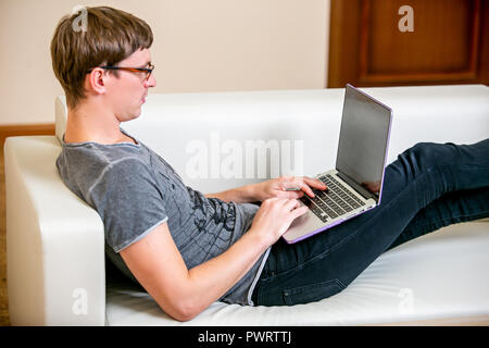 Concentrated young man with glasses working on a laptop in a home office. Prints on the keyboard and scrolls text on the screen while lying on the sofa Stock Photo