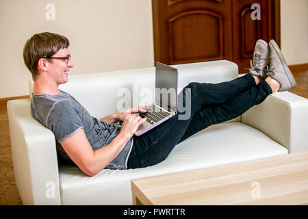 Concentrated young man with glasses working on a laptop in a home office. Prints on the keyboard, scans the text on the display and smiles while lying on the sofa Stock Photo