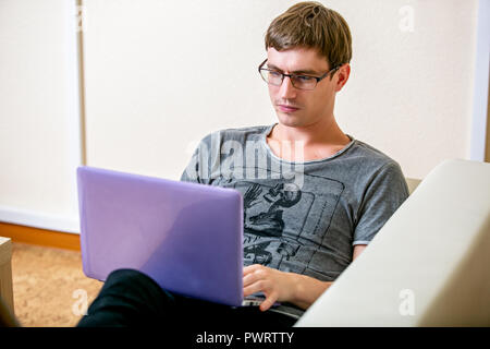 Concentrated young man with glasses working on a laptop in a home office. Prints on the keyboard and scans the text on the display, lying on the sofa Stock Photo