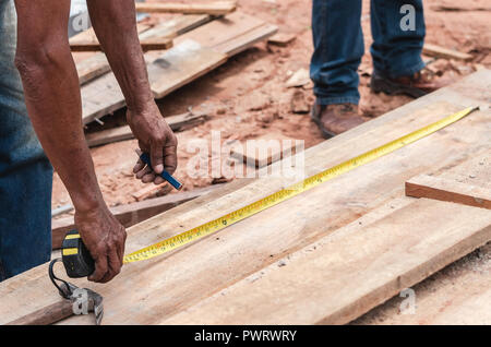 Hands of a carpenter with a measuring tape, measuring the size of a wooden plank to make a mark for cutting. Carpentry of a construction background. Stock Photo